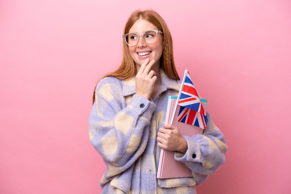 Molodaa ryzaa zensina s flagom soedinennogo korolevstva na rozovom fone smotrit vverh ulybaas (1)